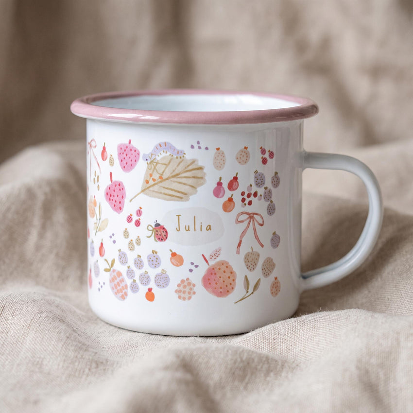 A white enamel mug with a pink rim and handle, featuring a colourful pattern of watercolour-style strawberries, berries, leaves, a ladybird, and a ribbon. The name Julia is printed in the centre within a light-coloured oval shape.