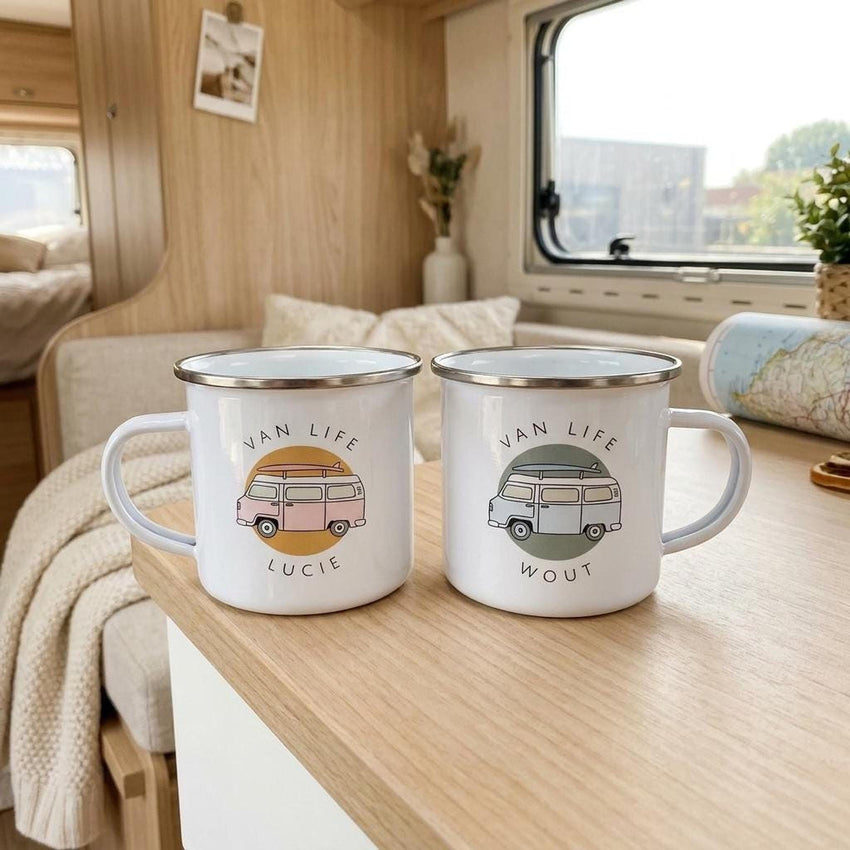 Two white enamel mugs with silver rims stand on a wooden table inside a campervan interior.