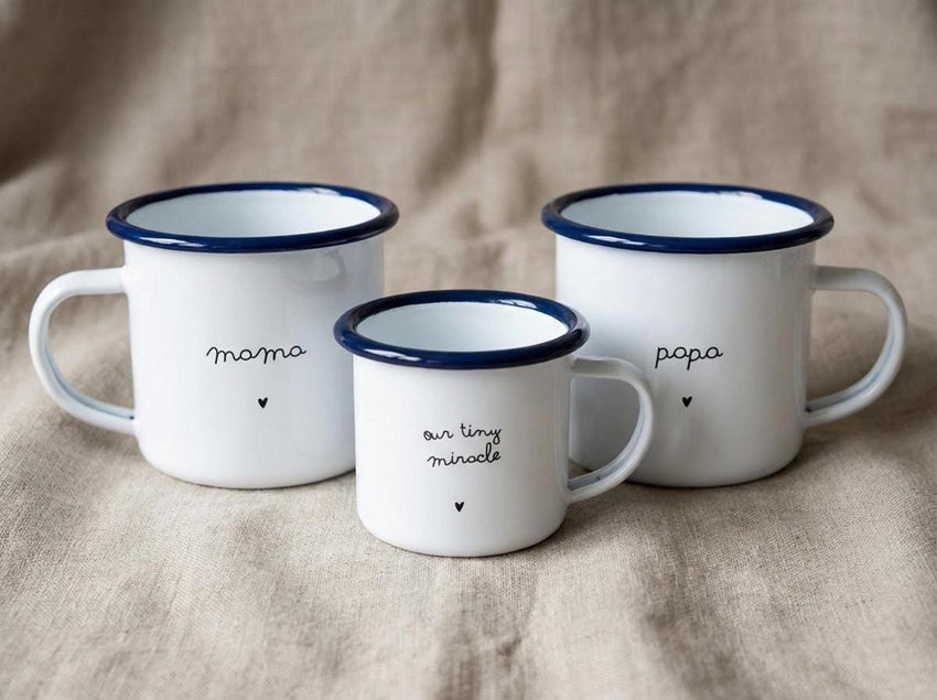 Three white enamel mugs with blue rims displaying mama, our tiny miracle, and papa with small hearts on a burlap background.