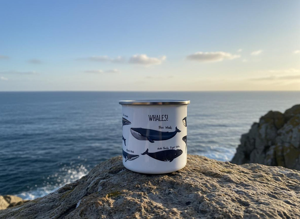 A white enamel mug with a silver rim featuring dark blue illustrations of various whale species and the text WHALES! printed in black. The mug displays labels for a Blue Whale and an Arctic Right Whale alongside other whale graphics.