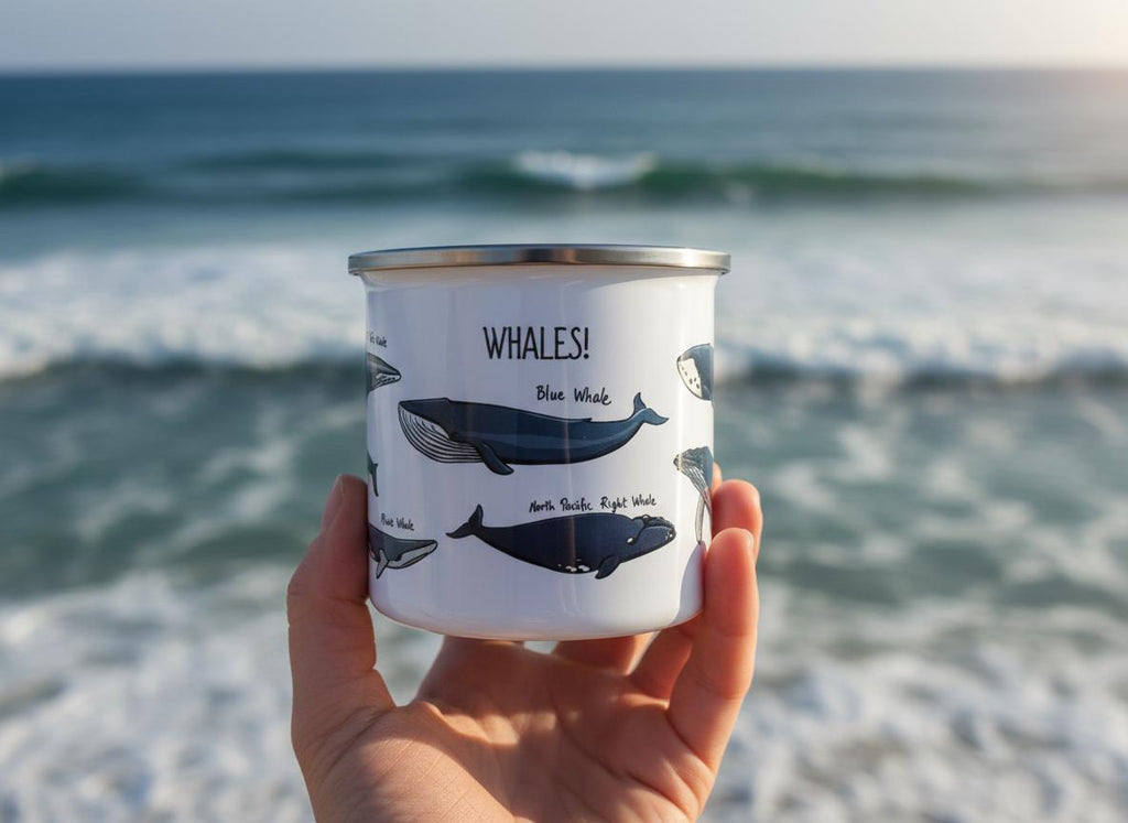 A white enamel mug with a silver rim featuring illustrations of various whale species and the text WHALES! in black lettering. The mug displays a Blue Whale and a North Pacific Right Whale with their names printed clearly below.