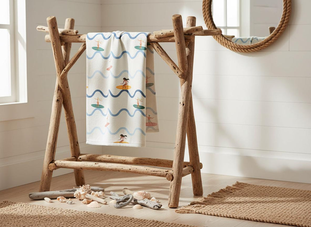 A cream-coloured towel featuring a repeating pattern of blue wavy lines and figures on surfboards, draped over a freestanding rack constructed from natural, unfinished driftwood branches with a sturdy, cross-legged frame design.