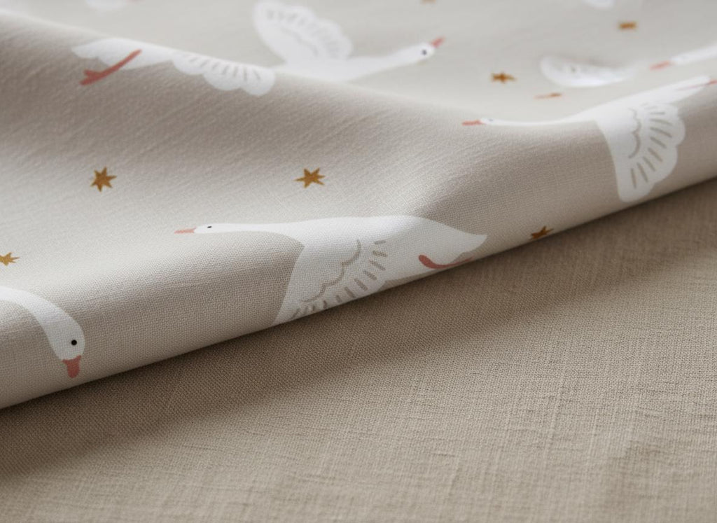 A light beige fabric featuring a repeating pattern of white geese in flight with orange beaks and feet, interspersed with small, dark orange four-pointed stars. The textile has a visible woven texture and is folded over itself.