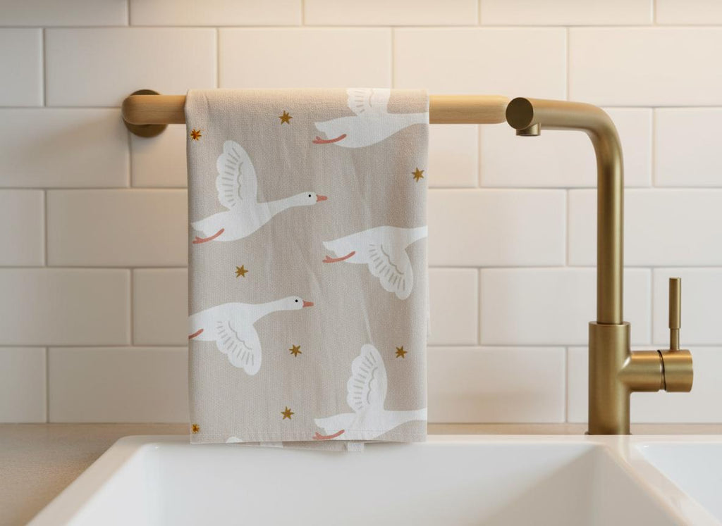 A beige kitchen towel featuring a repeating pattern of white flying geese with orange beaks and feet, interspersed with small gold stars. The towel is draped over a gold-toned metal bar attached to a white tiled wall above a sink.