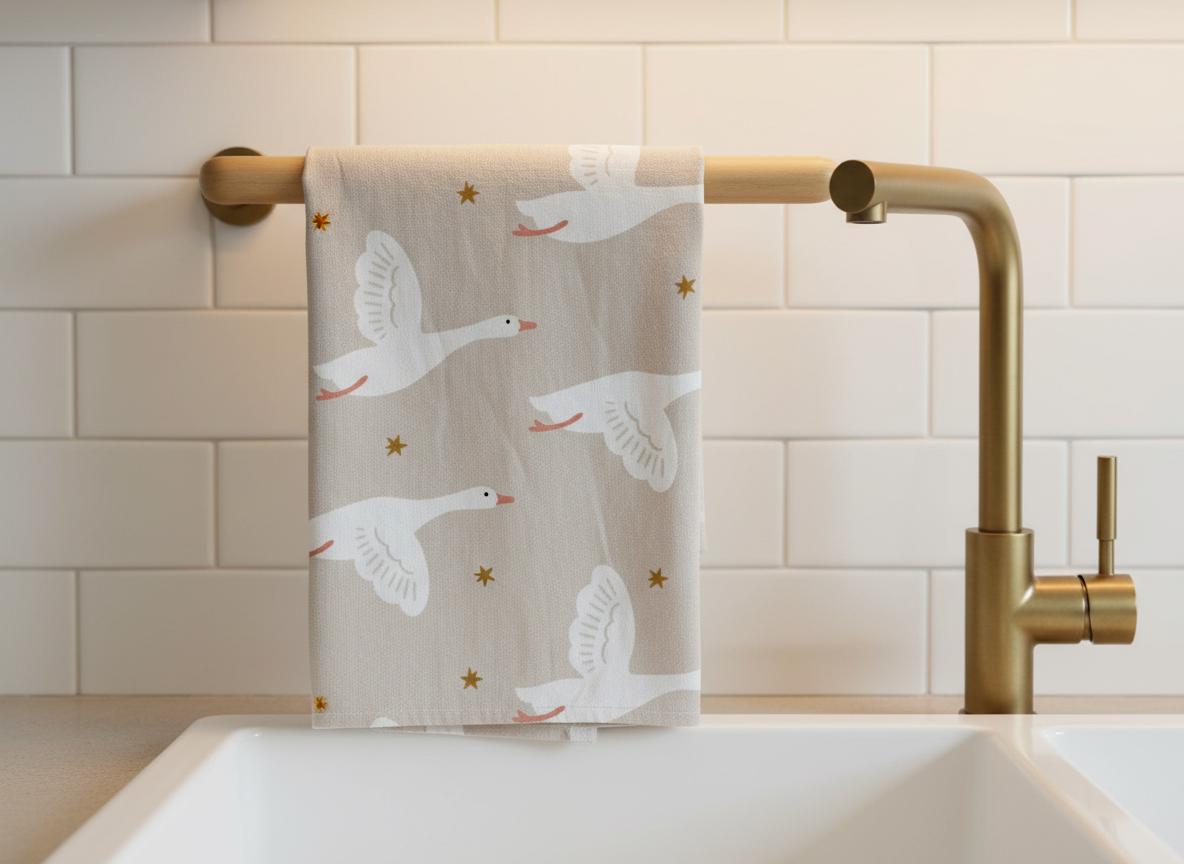 A beige kitchen towel featuring a repeating pattern of white flying geese with orange beaks and feet, interspersed with small gold stars. The towel is draped over a gold-toned metal bar attached to a white tiled wall above a sink.