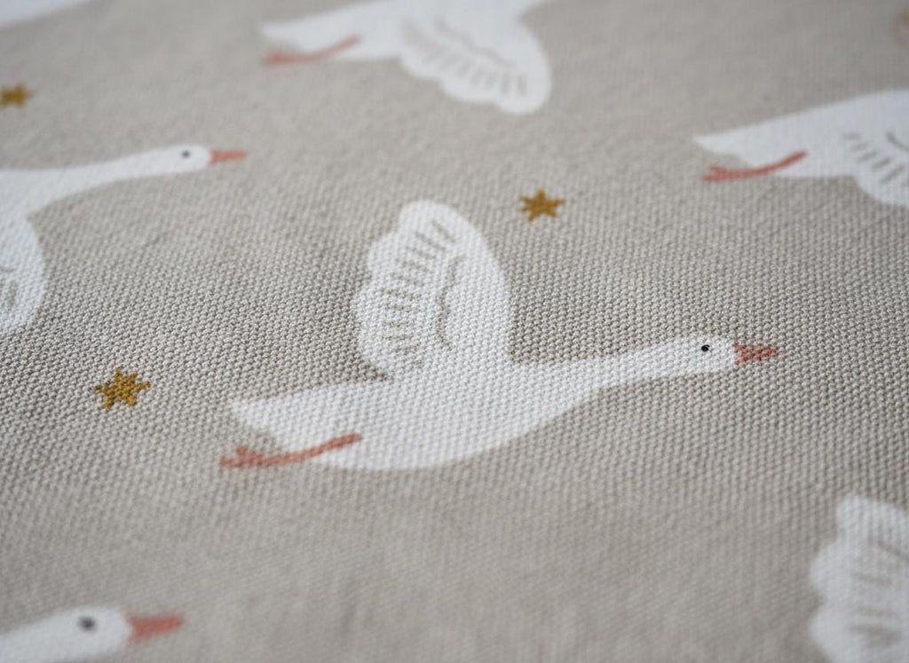 A light beige fabric featuring a repeating pattern of white flying geese with orange beaks and feet, interspersed with small, simple gold-coloured star shapes. The textile has a visible woven texture throughout the entire surface.