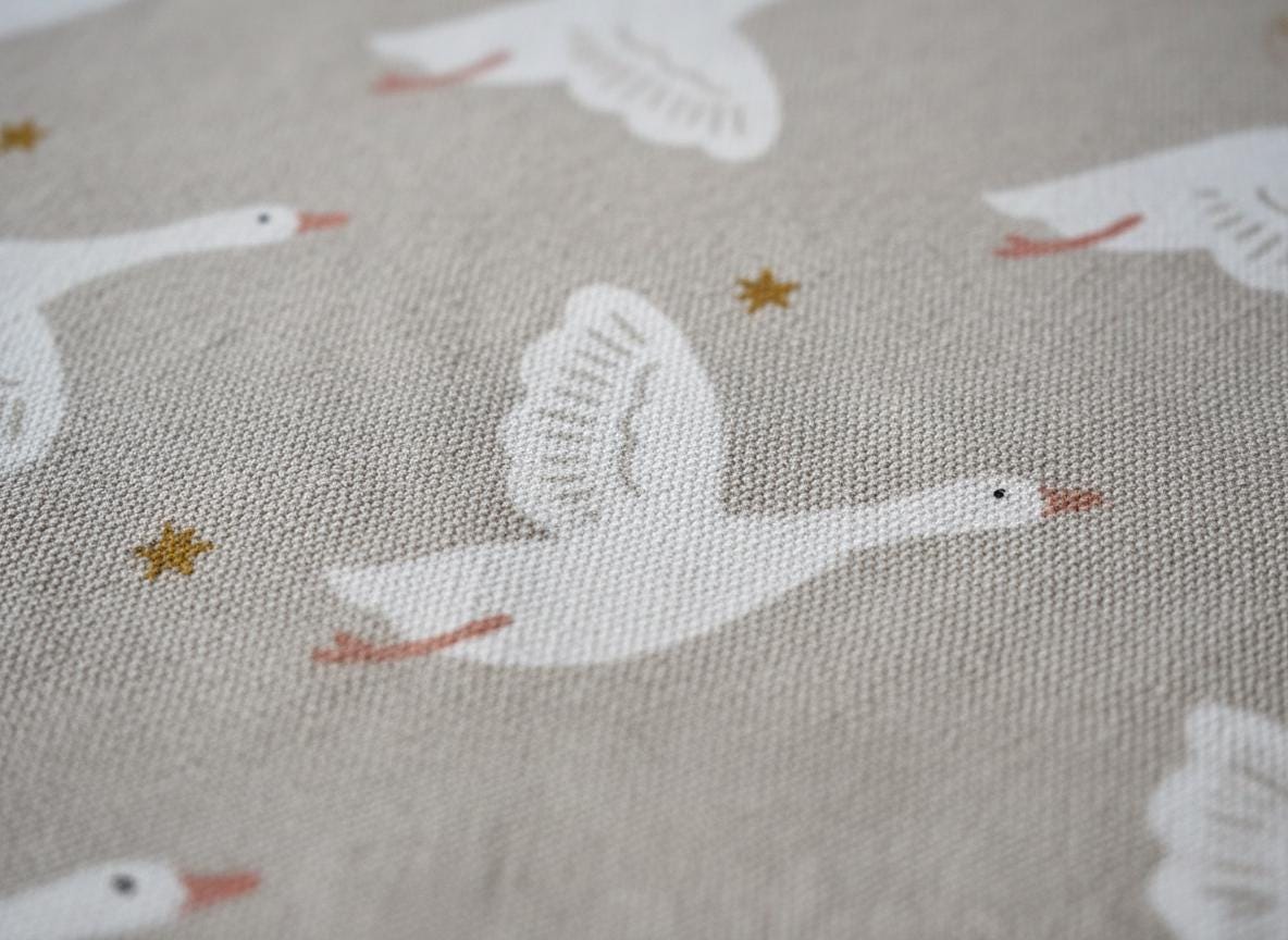 A light beige fabric featuring a repeating pattern of white flying geese with orange beaks and feet, interspersed with small, simple gold-coloured star shapes. The textile has a visible woven texture throughout the entire surface.