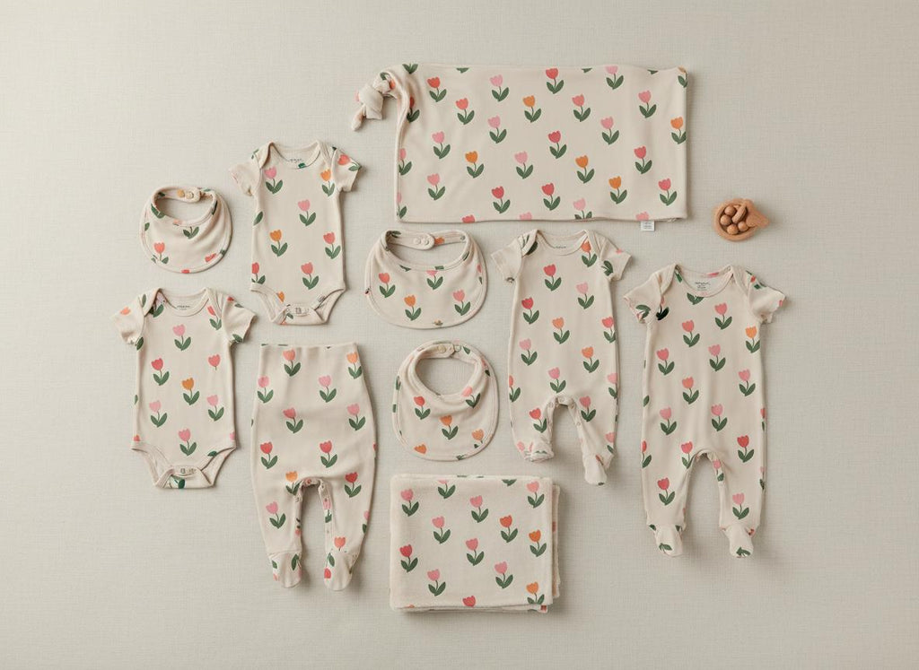 A collection of baby clothing and accessories in a cream colour featuring a repeating pattern of pink and orange tulips with green stems. The set includes three bodysuits, two pairs of footed trousers, three bibs, and two blankets.