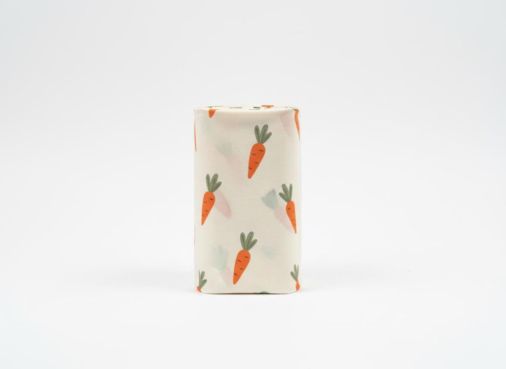 A cylindrical roll of off-white fabric featuring a repeating pattern of small orange carrots with green leafy tops. The fabric is tightly wound into a vertical tube shape, displaying the carrot print clearly across the entire surface.