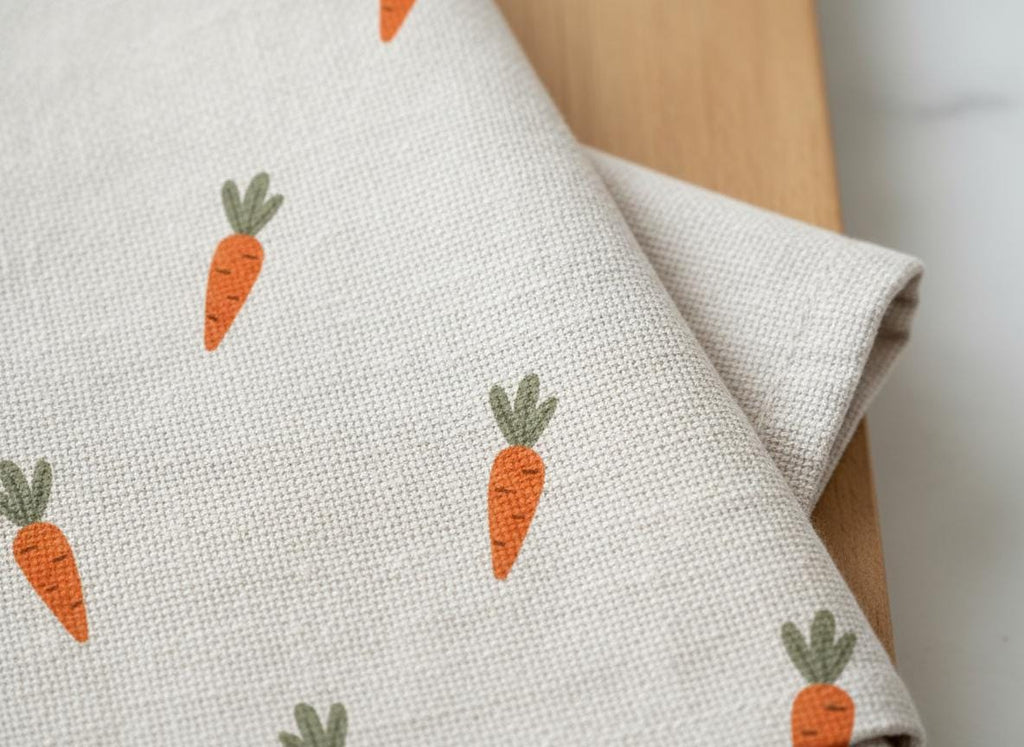 A light beige kitchen towel featuring a repeating pattern of small orange carrots with green leafy tops. The fabric has a visible woven texture, and the carrots are evenly spaced across the surface of the folded textile material.