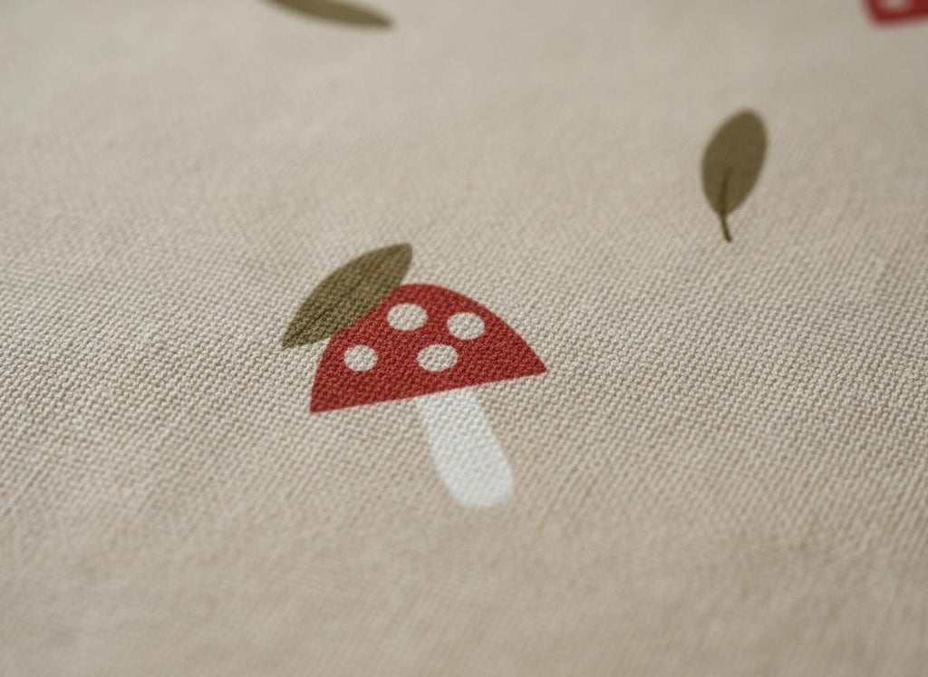 A close-up of a beige fabric featuring a repeating pattern of red mushrooms with white spots and green leaves. The fabric has a textured appearance, possibly linen or cotton.