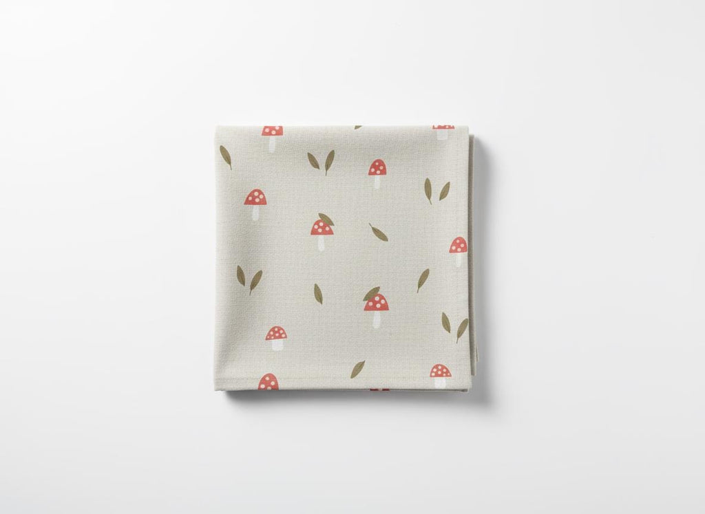 Folded square fabric featuring a pattern of red and white mushrooms and brown leaves on a light beige background. The fabric is likely cotton or linen.