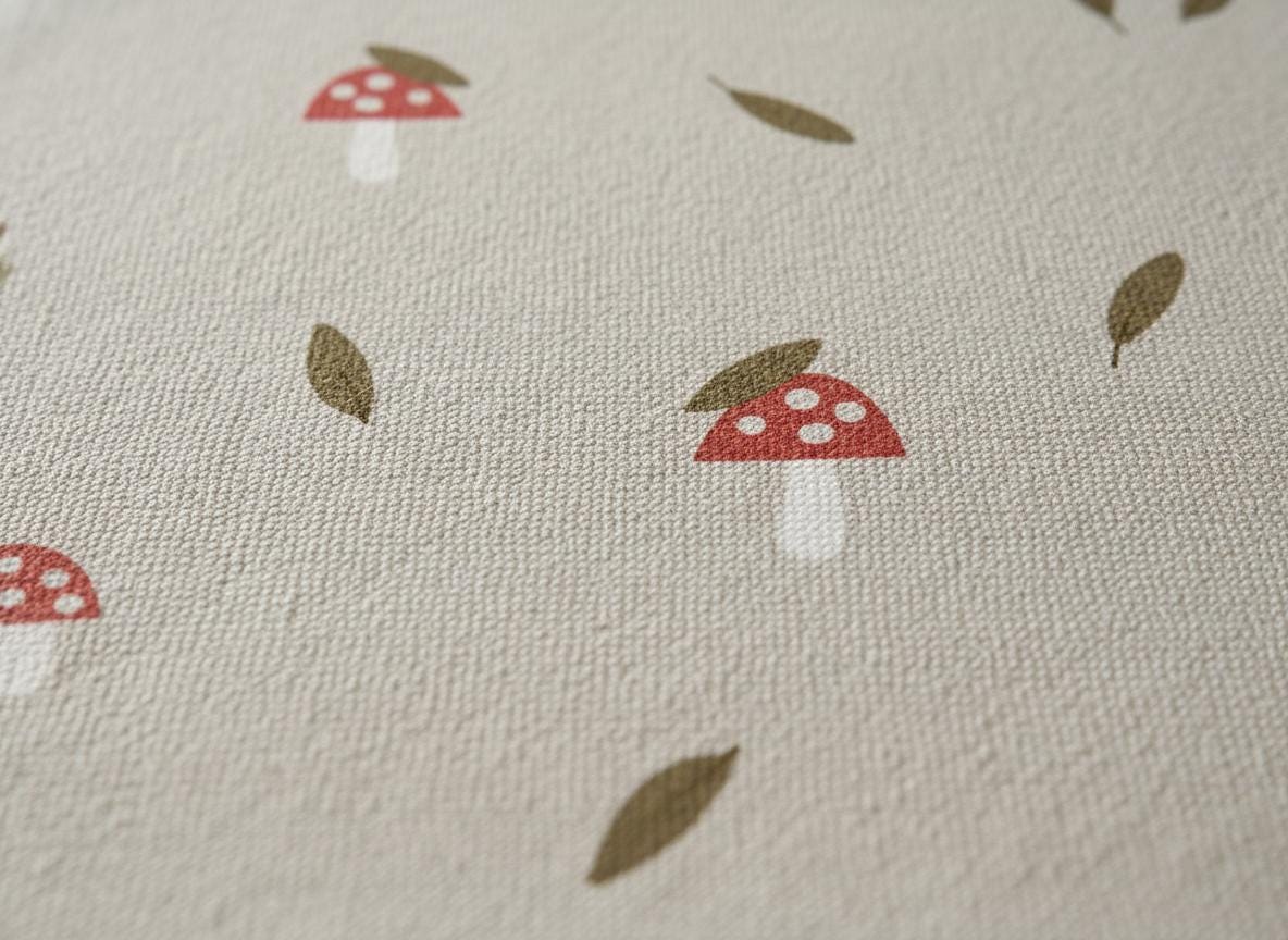Close-up of a beige fabric featuring a repeating pattern of red mushrooms with white spots and brown leaves. The fabric has a textured appearance, possibly linen or cotton.