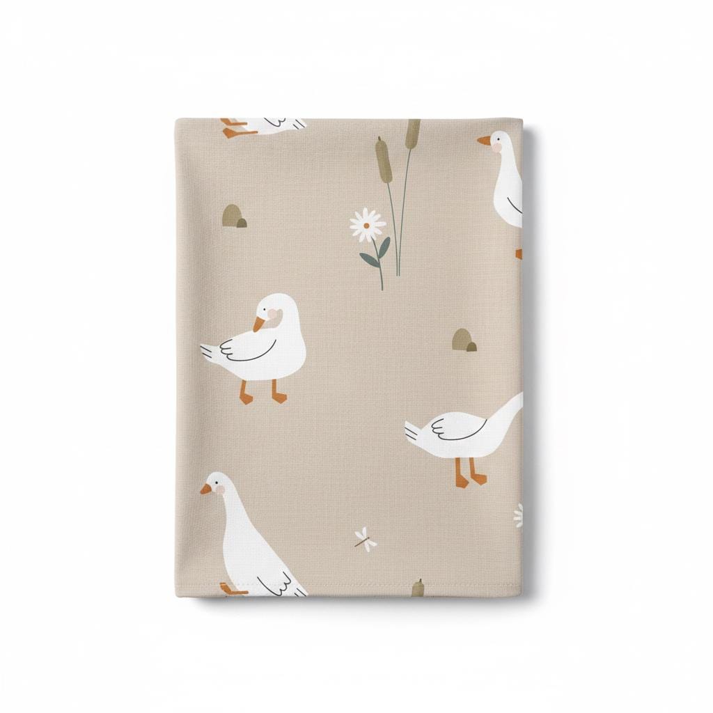 A light beige fabric featuring a repeating pattern of white geese with orange beaks and feet, interspersed with small white daisies, brown cattails, tiny dragonflies, and small grey stones scattered across the surface.