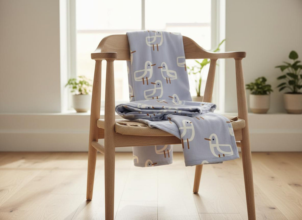 Folded blanket in the lavender-blue Seagull pattern draped over a wooden chair in a bright room with plants.