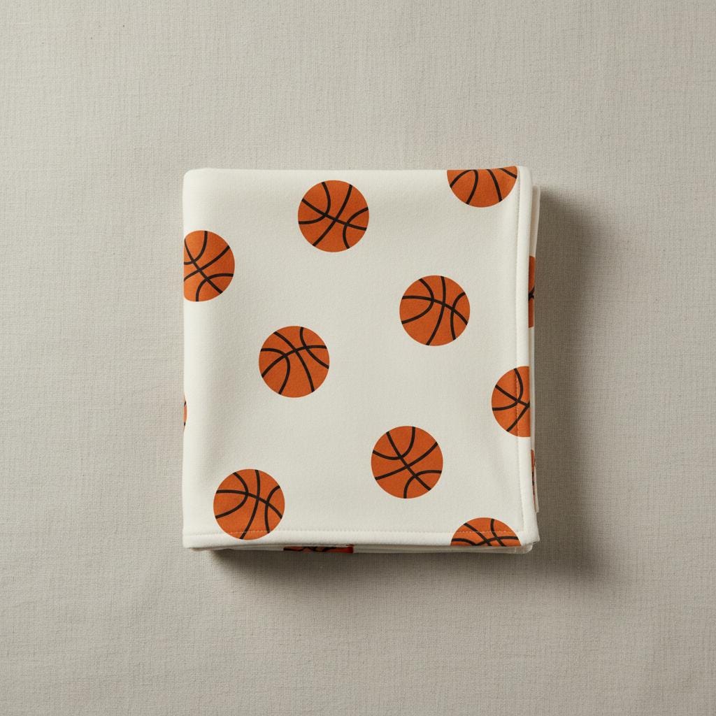Folded cream blanket printed with the Basketball pattern of orange basketballs, on a grey background.