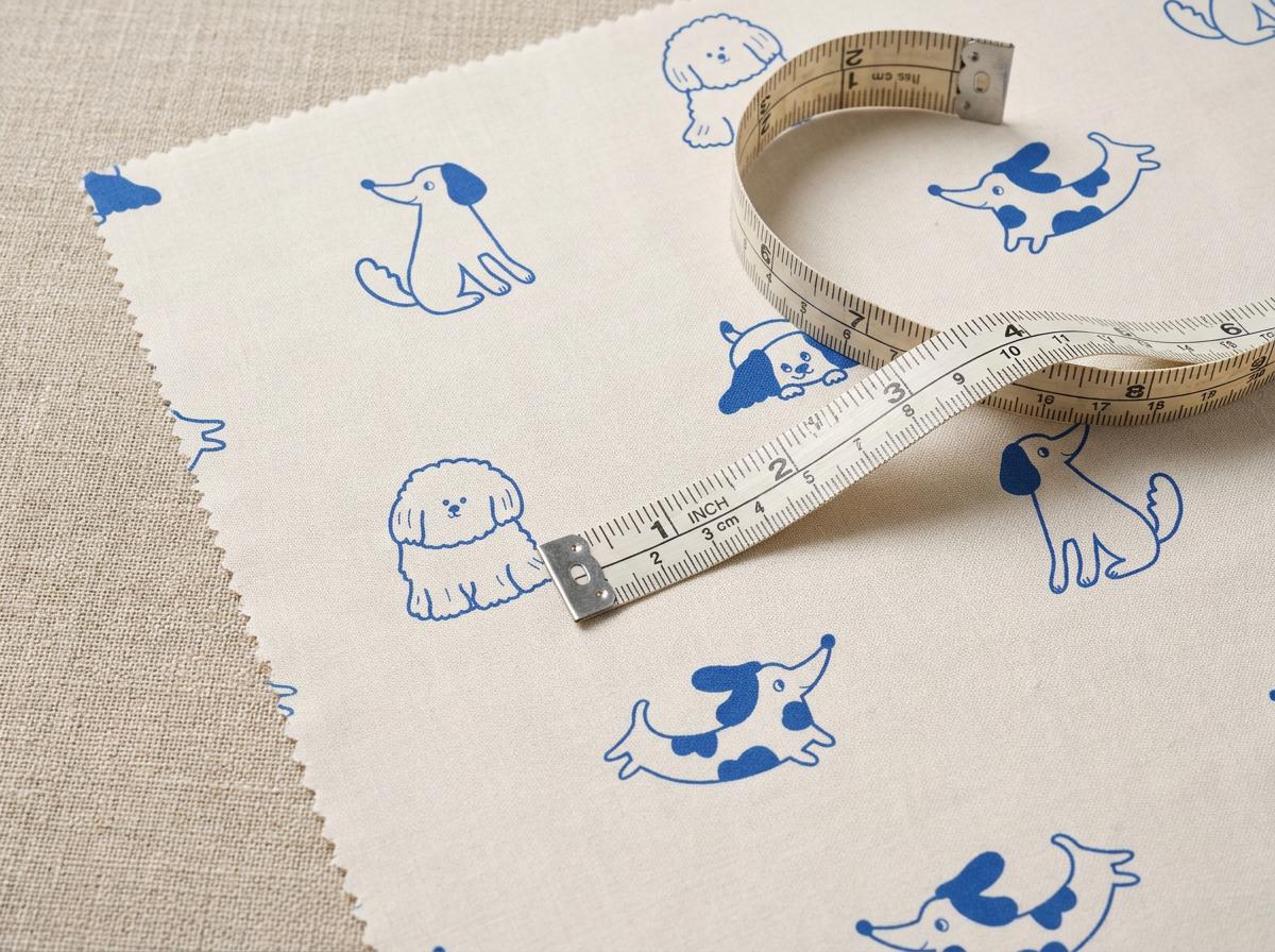 A piece of off-white fabric featuring a repeating pattern of blue line-art dogs. A flexible measuring tape is placed on top, showing markings in inches and centimetres, with the tape indicating a length of approximately 10 centimetres.
