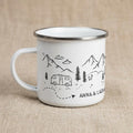 A white enamel mug with a silver rim and a black line art illustration featuring mountains, pine trees, a camper van, and a dotted path. The text ANNA & LUCA is printed in black on the side of the mug, which has a single handle.