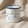 A white enamel mug with a dark blue rim and handle, featuring a repeating pattern of light blue wavy lines and small illustrations of people surfing on colourful surfboards in various positions across the exterior surface.