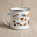 A white enamel mug with a silver rim and handle, featuring a detailed botanical illustration of various mushrooms including King Trumpet, Boletus, and Shiitake, along with pine needles, acorns, and oak leaves with text labels.