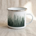 Forest enamel mug: white camping mug with a silver rim, wrapped in a misty landscape of dark green pine trees fading upwards.