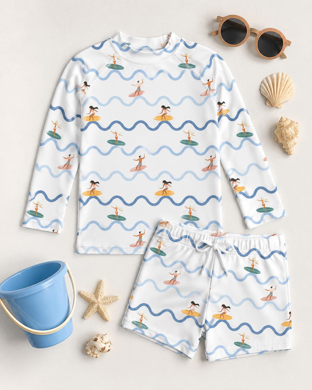 A white long-sleeve rash guard and matching shorts set featuring a repeating pattern of blue wavy lines and small figures surfing on colourful surfboards. The set is displayed alongside brown sunglasses, seashells, a starfish, and a blue bucket.