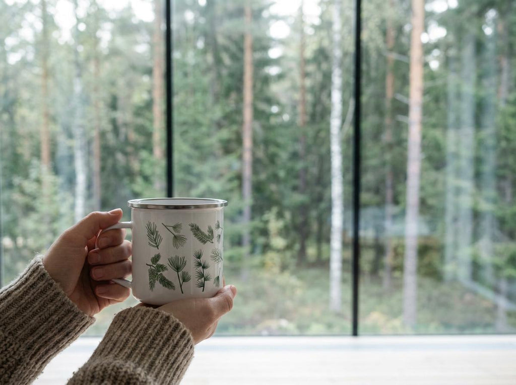 A white enamel mug with a silver rim featuring a green botanical pattern of various pine needles and branches. The mug has a cylindrical body and a side handle, held by hands wearing a beige ribbed knit sweater sleeve.