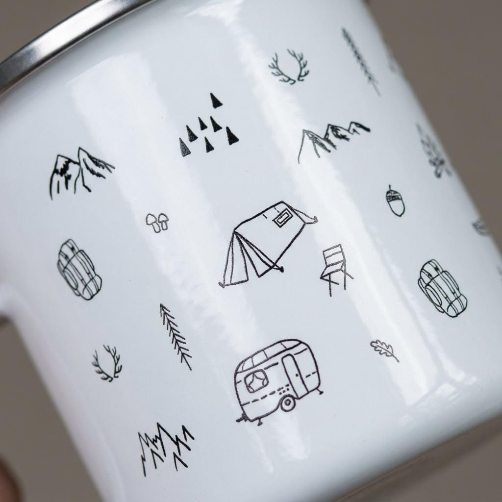 Close-up of the Camping mug design: black line-art tent, vintage caravan, mountains, backpacks and camp chair on white enamel.