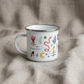 Nordic Folk Art enamel mug on soft grey linen, decorated with a folk-style snake, flowers, ferns and a crescent moon in red, mustard and green.
