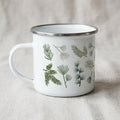 A white enamel mug with a silver-toned rim and a handle on the left side. The exterior features a repeating pattern of various green pine needles and evergreen branch illustrations arranged across the surface of the white metal cup.
