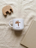 A white cup featuring a brown giraffe illustration and the name MILAN printed in black text. The cup has a rounded handle and a smooth finish, positioned near a wooden elephant figurine and a closed book with a tan cover.