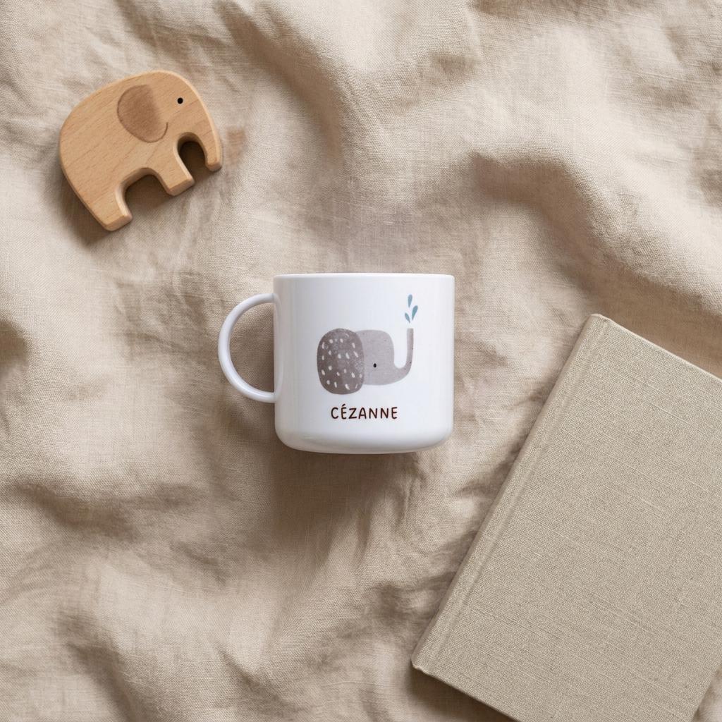 A white cup featuring a grey elephant illustration with a blue water spray and the name CÉZANNE printed in brown text, accompanied by a light brown wooden elephant figurine and a closed book with a light beige cover.