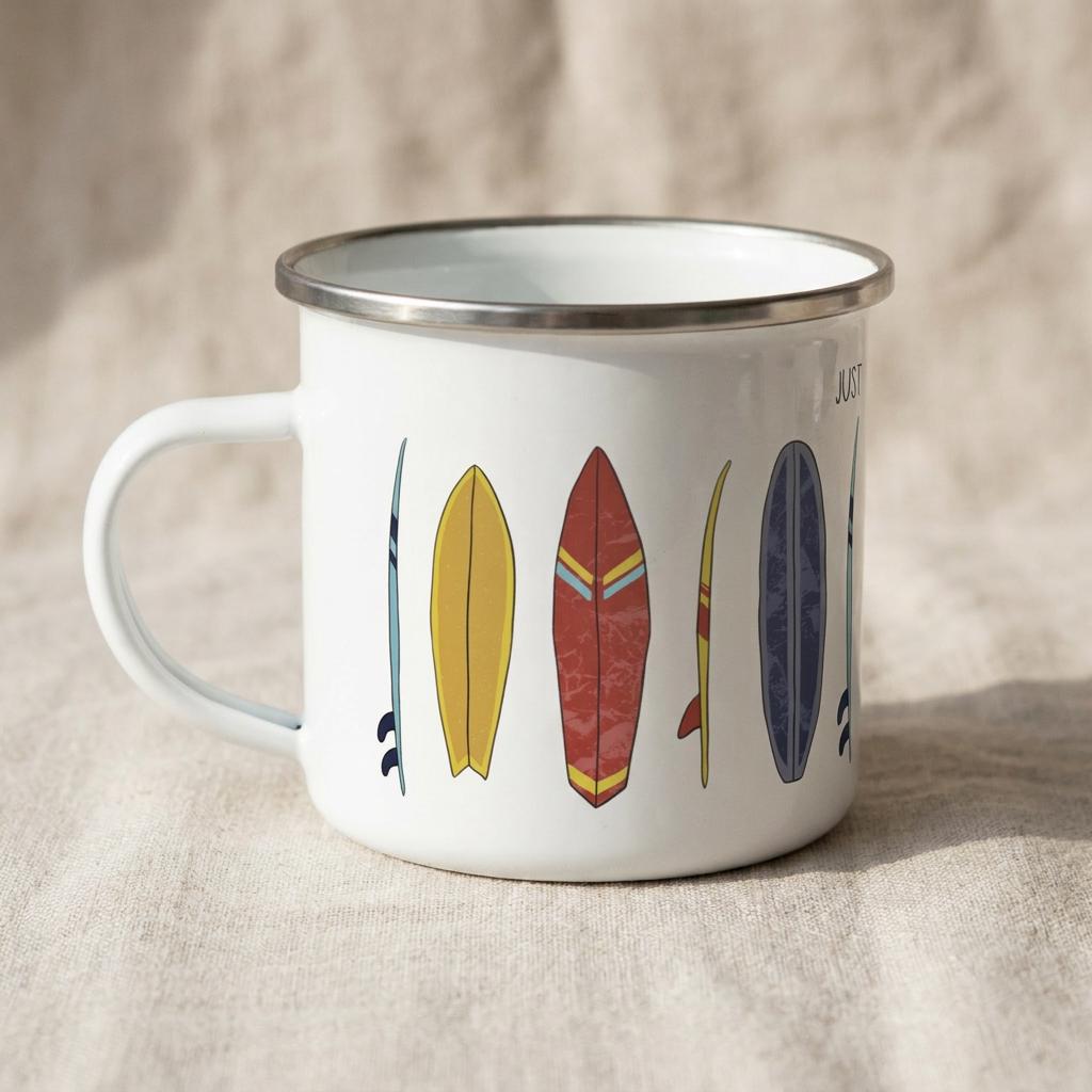 A white enamel mug with a silver-toned rim and a handle, featuring a graphic print of various colourful surfboards in shades of yellow, red, and blue, along with the partial text "JUST" visible on the side of the mug.