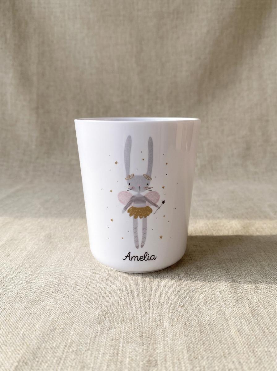 A white tumbler featuring a graphic illustration of a grey rabbit fairy with pink wings, a gold skirt, and a wand. The design includes small gold stars scattered around the rabbit, and the name Amelia is printed in black script below.