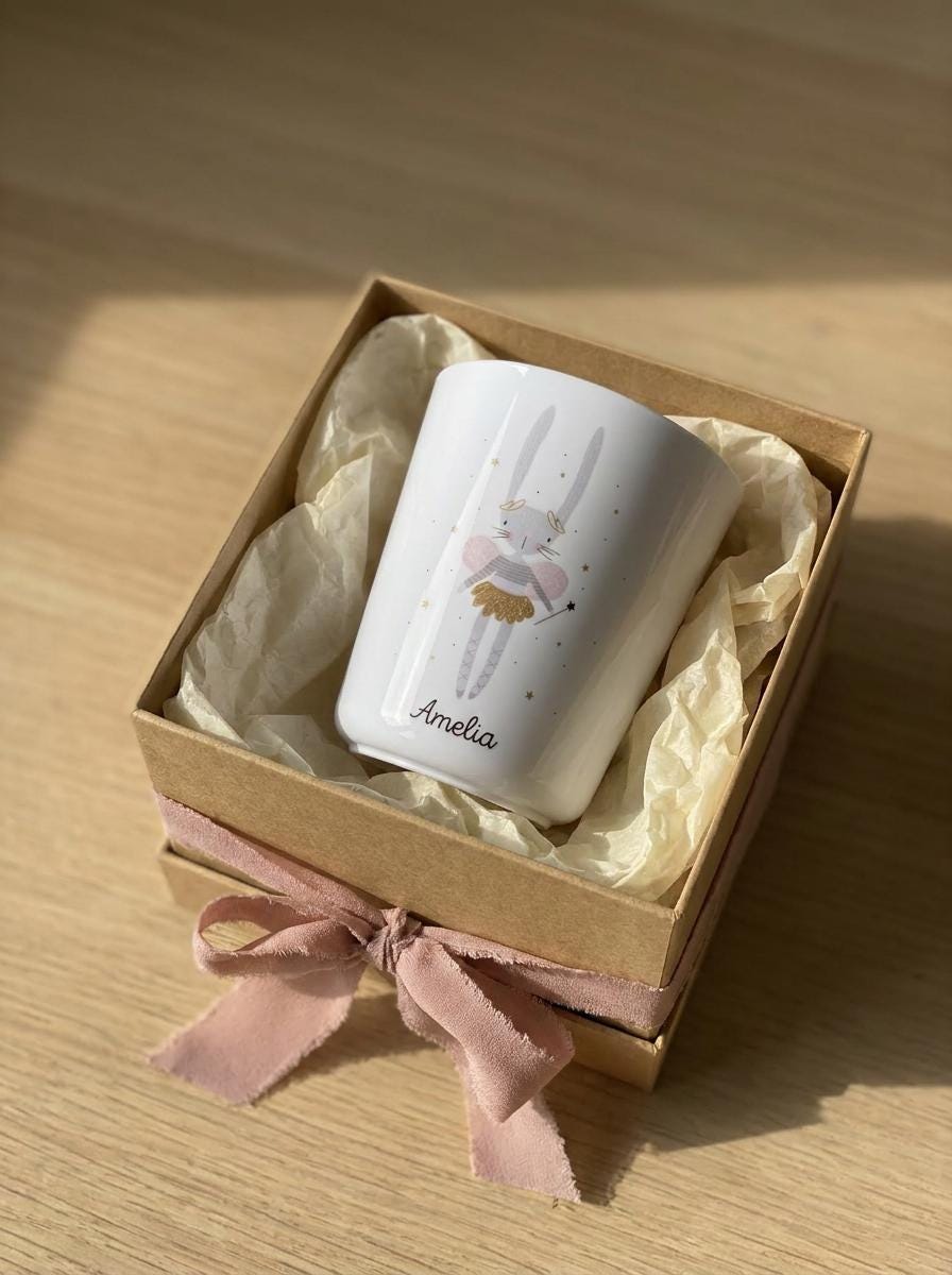 A white tumbler featuring a graphic of a rabbit with wings and a gold skirt, personalised with the name Amelia in black script, nestled inside a square brown cardboard gift box lined with cream tissue paper and a pink ribbon.