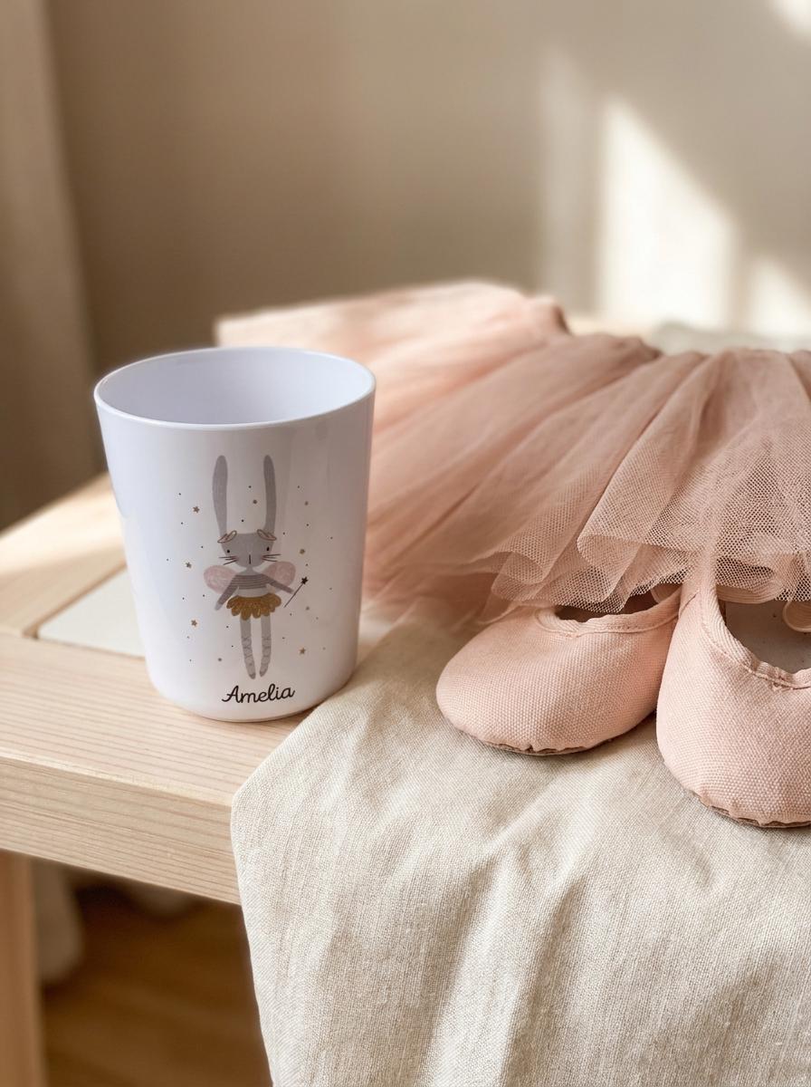 Personalised Bunny Ballerina cup with the name Amelia: white handleless kids cup next to pink ballet shoes and a tulle tutu on a wooden stool.
