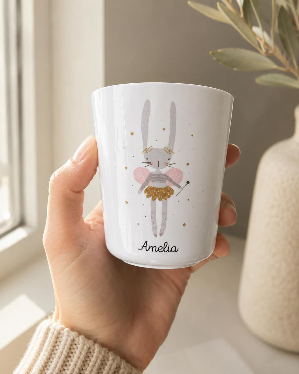 A white tumbler featuring a grey rabbit illustration with pink wings, a gold skirt, and a gold headband. The rabbit holds a small black wand, surrounded by gold stars. The name Amelia is printed in black script at the bottom of the cup.