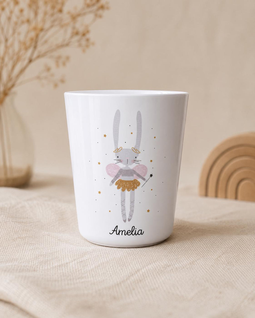A white cylindrical cup featuring a printed illustration of a grey rabbit fairy with pink wings, a gold skirt, and a wand. The rabbit is surrounded by small gold stars. The name Amelia is printed in black script lettering at the bottom.
