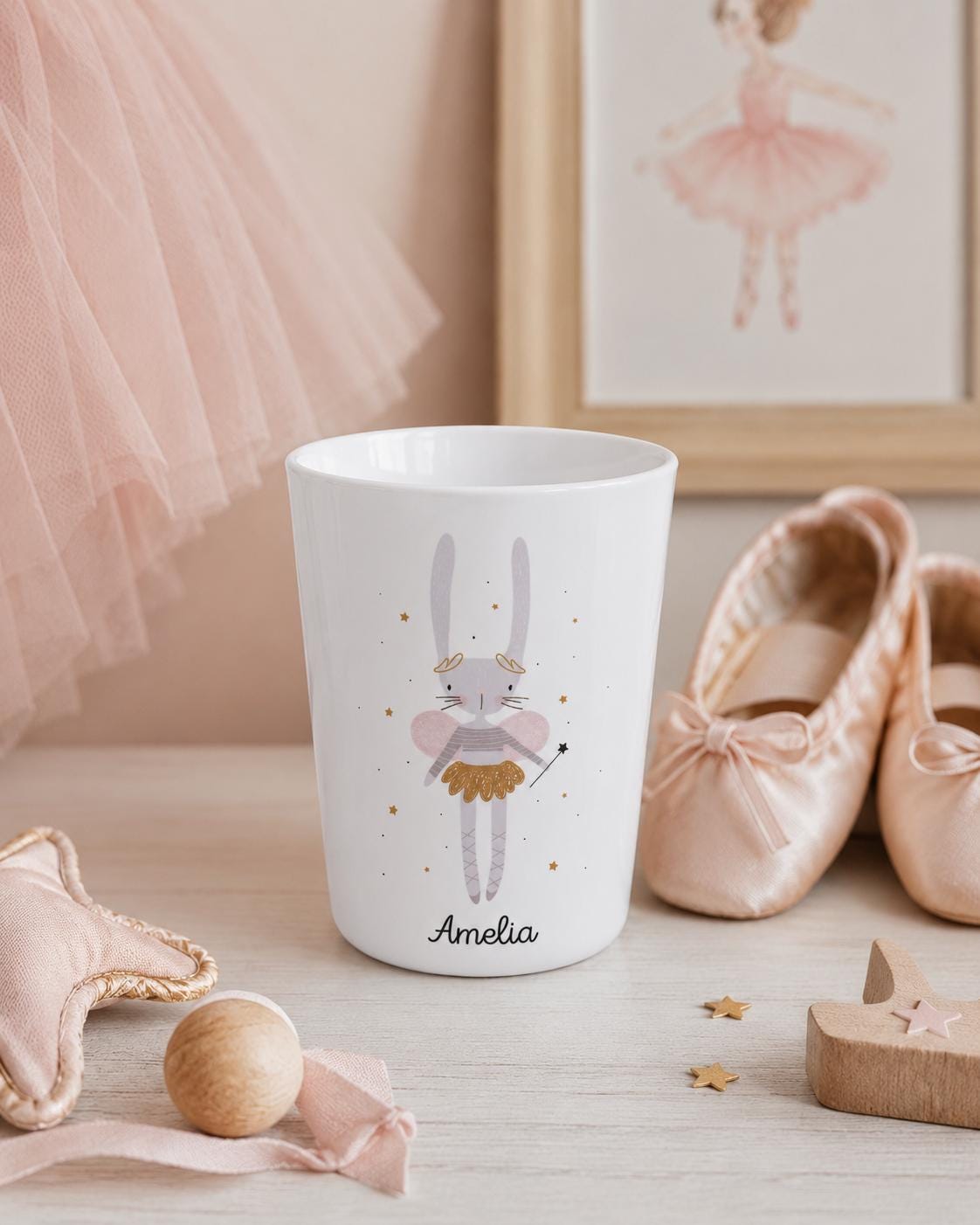 A white cylindrical cup featuring a printed illustration of a grey rabbit fairy with pink wings, a gold skirt, and a wand. The name Amelia is printed in black script below the rabbit, surrounded by small gold stars on the cup surface.