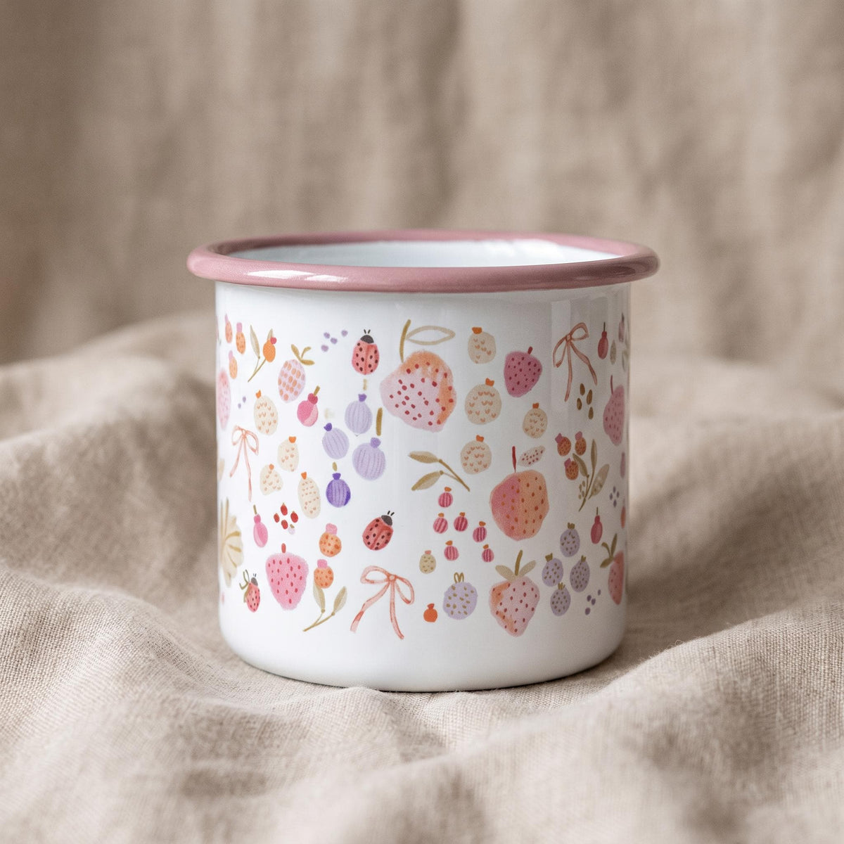 A white enamel mug with a pink rim featuring a repeating pattern of small pink and purple fruits, berries, ladybugs, and light pink bows scattered across the exterior surface. The mug has a cylindrical shape and a smooth finish.