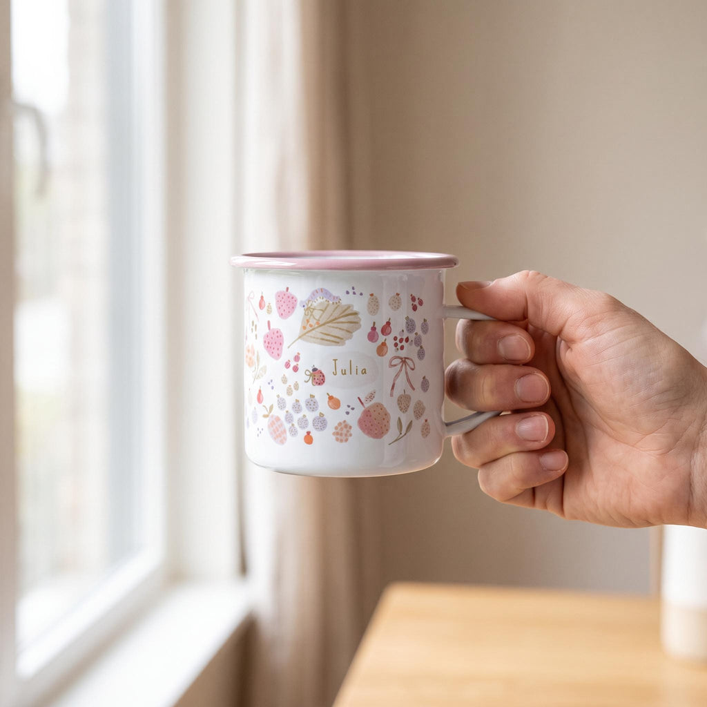 A white enamel mug with a pink rim and handle, featuring a pattern of small colourful fruits, leaves, and a ladybird. The name Julia is printed in the centre of the design within a small oval shape on the side of the mug.