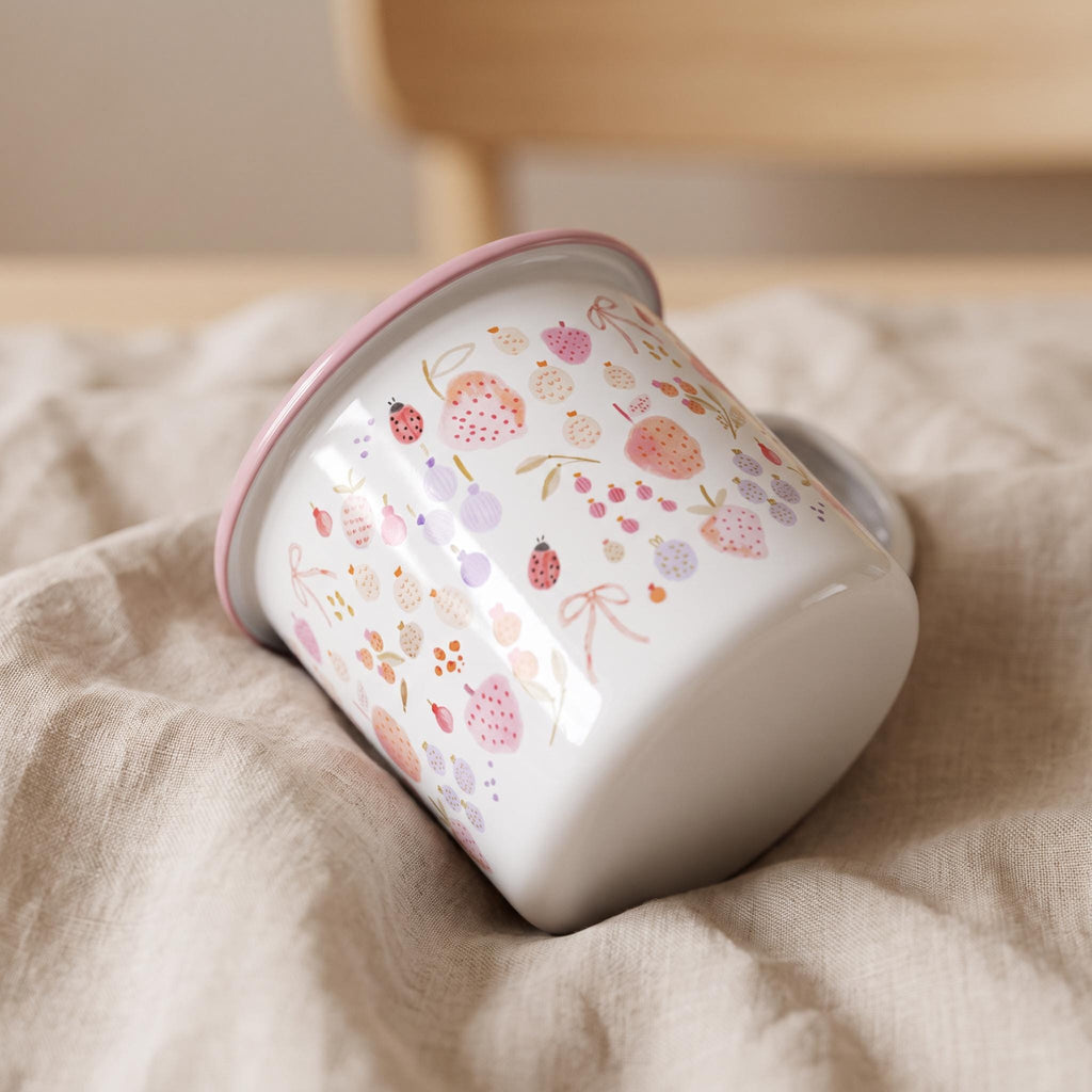 A white enamel mug with a pink rim featuring a repeating pattern of small pink and purple fruits, berries, bows, and ladybugs. The mug is tilted on its side, showcasing the cylindrical body and the smooth, glossy finish of the exterior.