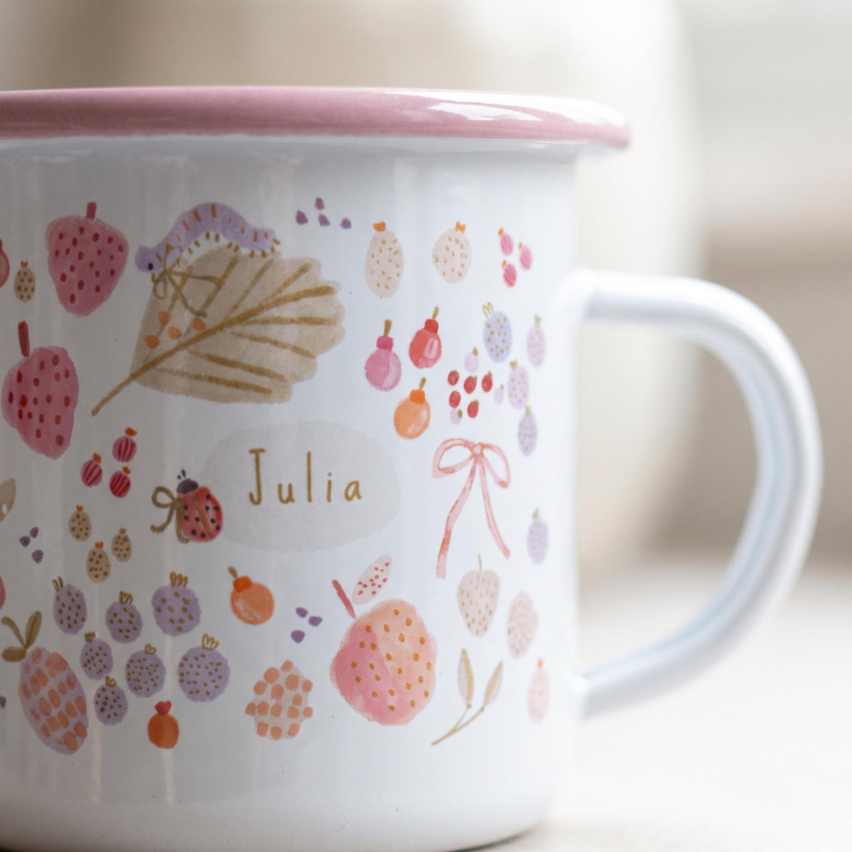 A white enamel mug with a pink rim and handle, featuring a colourful pattern of small fruits, berries, a ladybug, a caterpillar, and a bow. The name Julia is printed in the centre on a light oval background in a simple font.