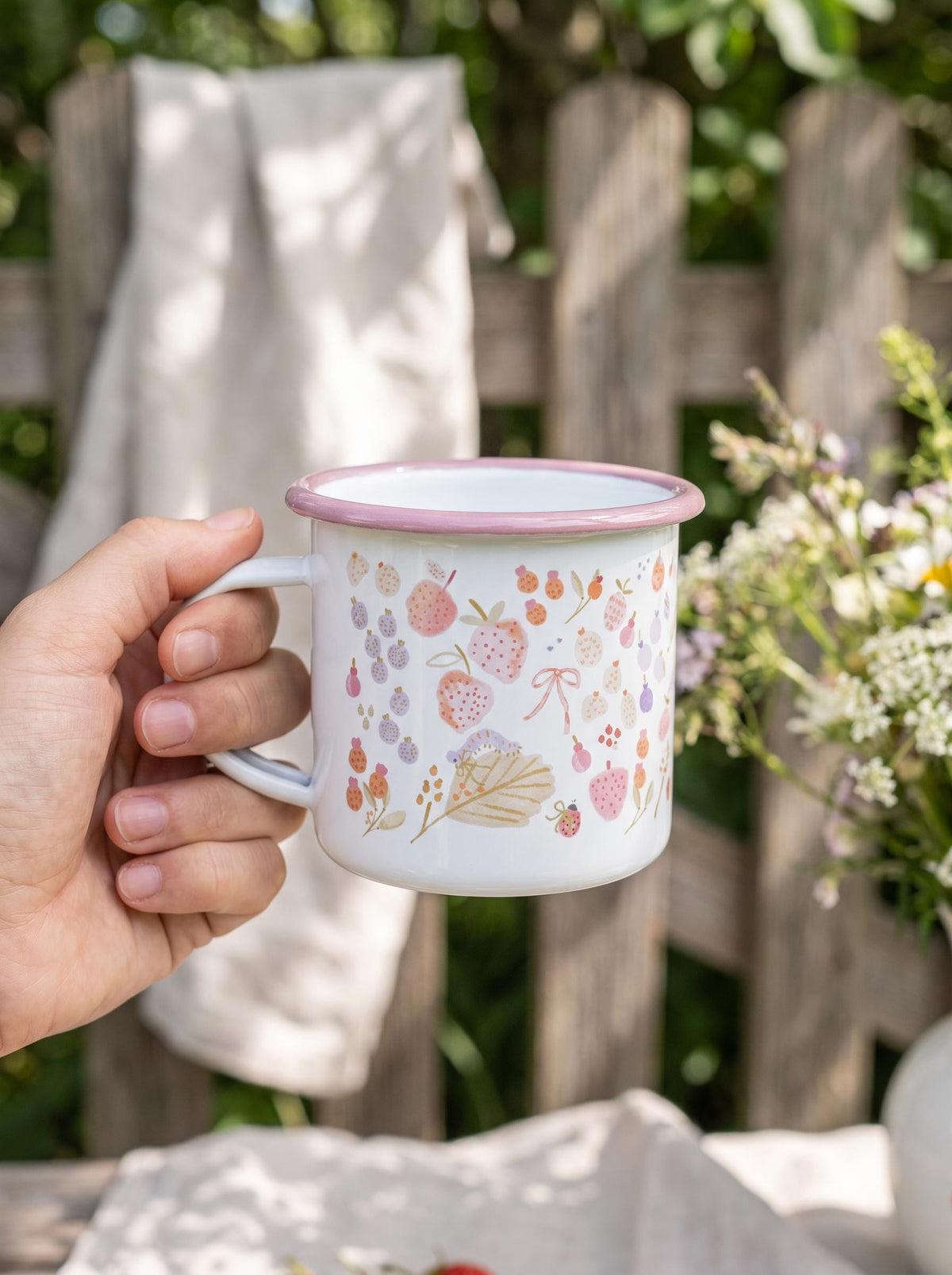 A white enamel mug with a pink rim featuring a delicate pattern of strawberries, small flowers, leaves, and a tiny ladybug. The mug has a side handle and a smooth, glossy finish, showcasing a light-coloured botanical illustration.