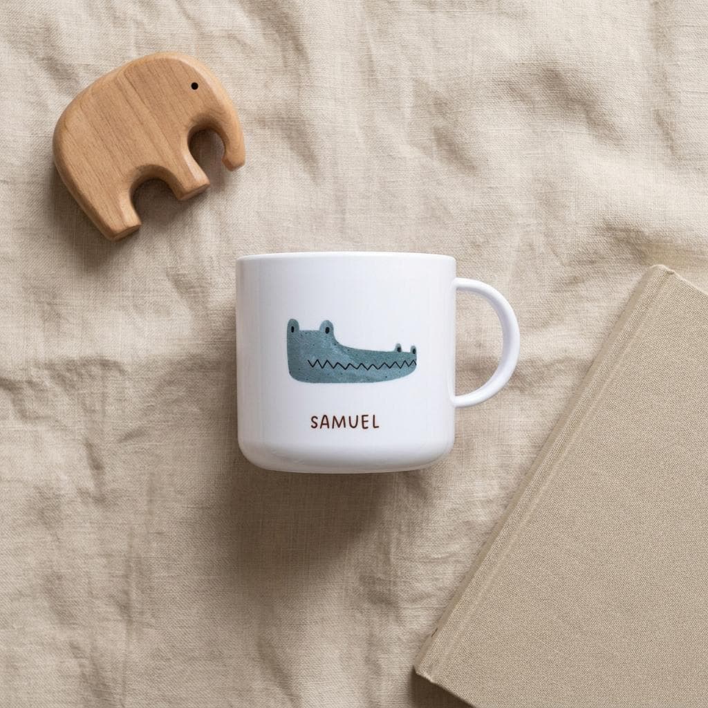 A white kids cup featuring a blue illustrated crocodile and the name SAMUEL printed in brown letters. Beside the cup is a light brown wooden elephant figurine and a closed beige book with a textured cover.