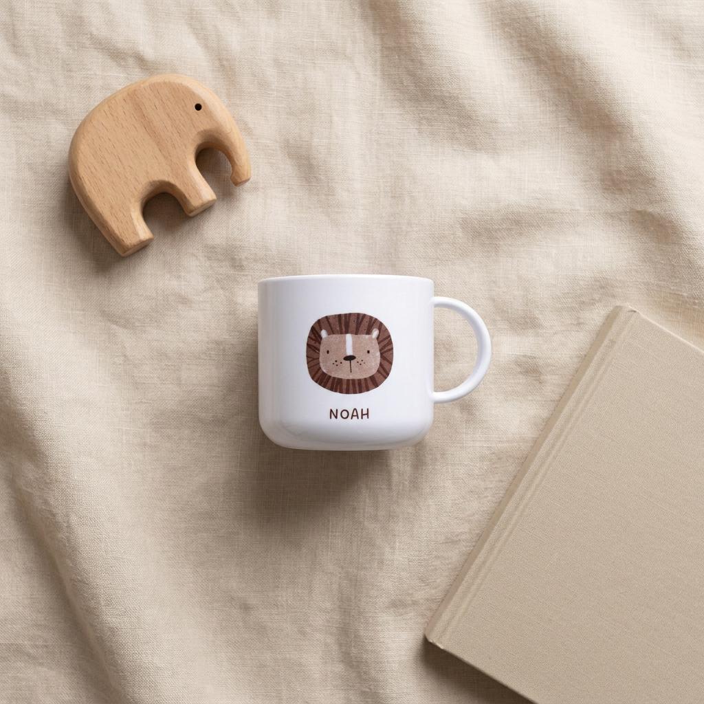 A white kids cup featuring a brown lion illustration and the name NOAH printed in brown letters, accompanied by a light brown wooden elephant figurine and a closed beige book with a hard cover and rounded corners.