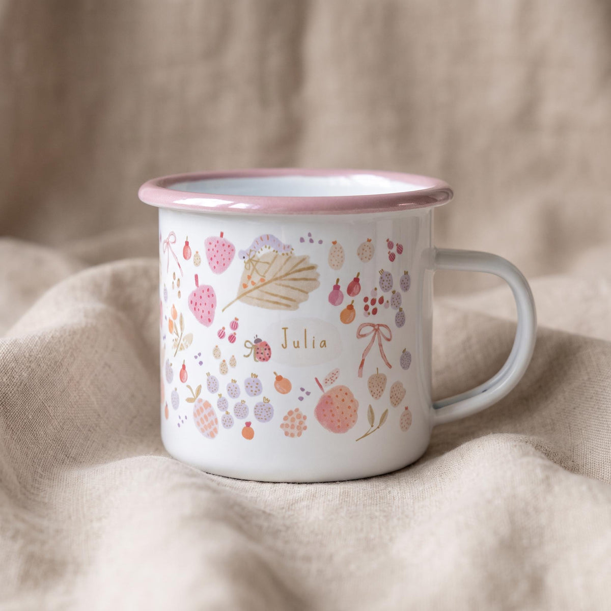 A white enamel mug with a pink rim and handle, featuring a colourful pattern of small fruits, leaves, bows, and a ladybird. The name Julia is printed in the centre within a light oval shape, surrounded by delicate watercolour-style illustrations.