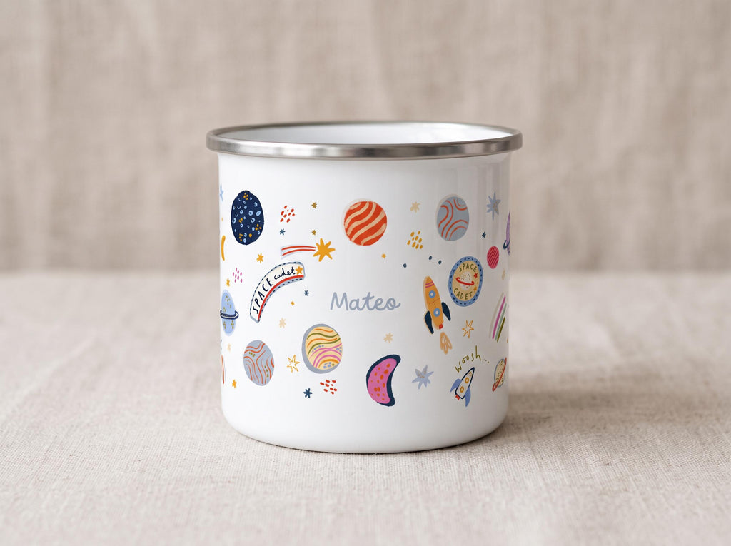 A white enamel mug with a silver rim featuring a colourful space-themed pattern of planets, stars, rockets, and the text "Space cadet" and "Mateo" printed on the side. The design includes various celestial bodies in shades of orange, blue, and pink.