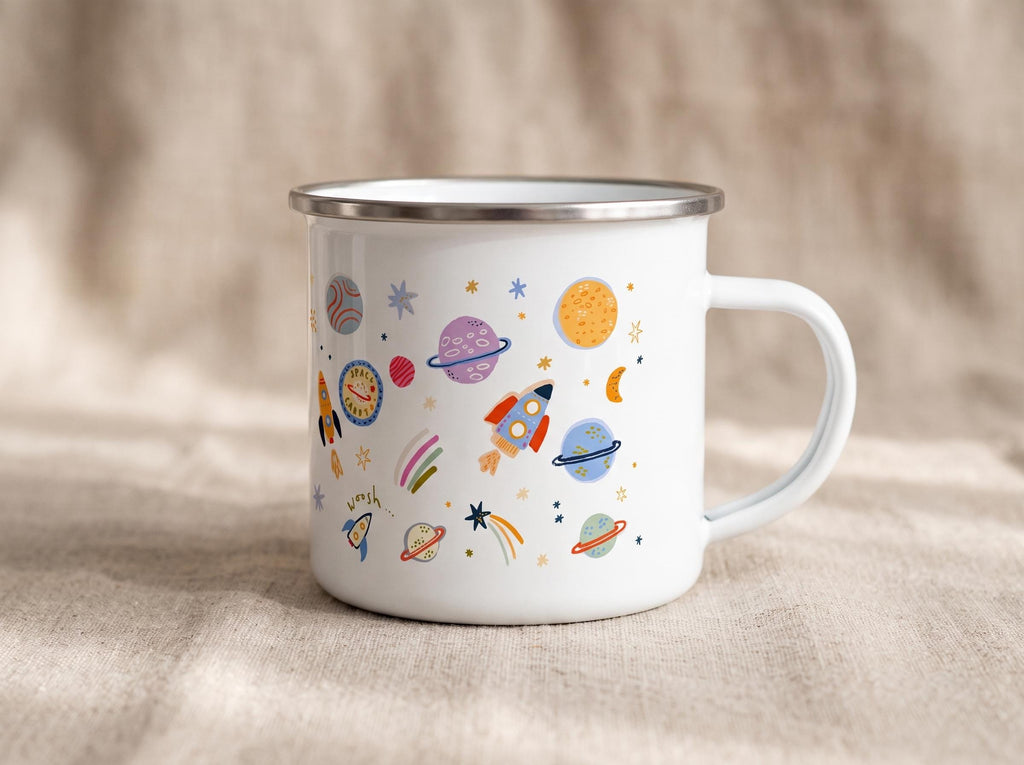 A white enamel mug with a silver rim and a handle, featuring a colourful pattern of space-themed illustrations including rockets, various planets with rings, stars, a crescent moon, and the word woosh written in small cursive script.