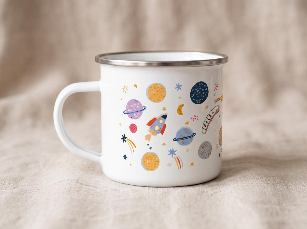 A white enamel mug with a silver rim and a handle, featuring a colourful space-themed pattern of planets, stars, a rocket ship, and a banner with the text SPACE AWAITS in a playful font, set against a neutral-toned fabric background.